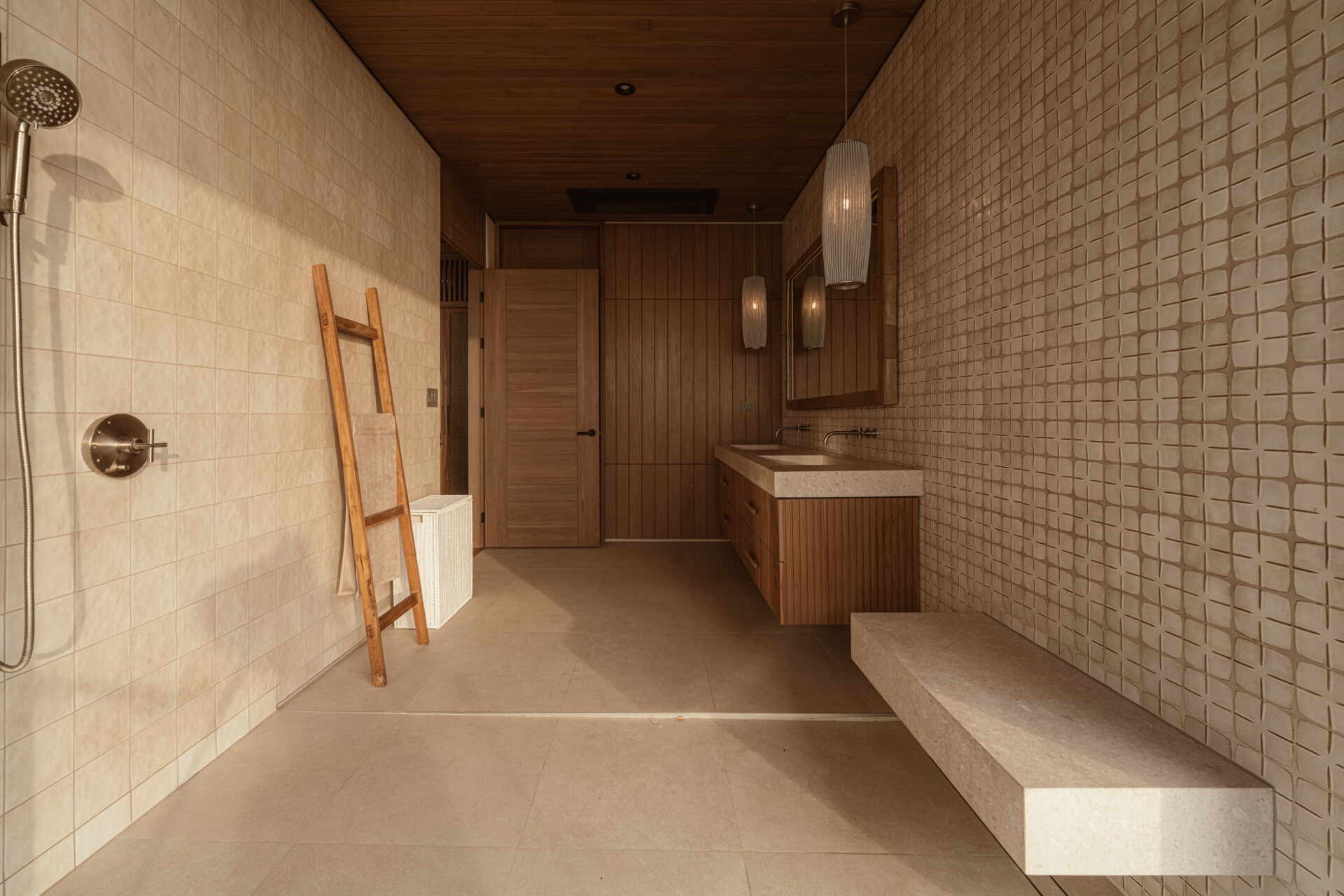This ensuite bathroom continues this calming palette with smooth stone surfaces, warm wood paneling, and softly patterned wall tiles. A floating bench adds function without heaviness, while sculptural pendant lighting and a simple wood-framed mirror elevate the space just enough, and folding glass doors open to the outdoors.