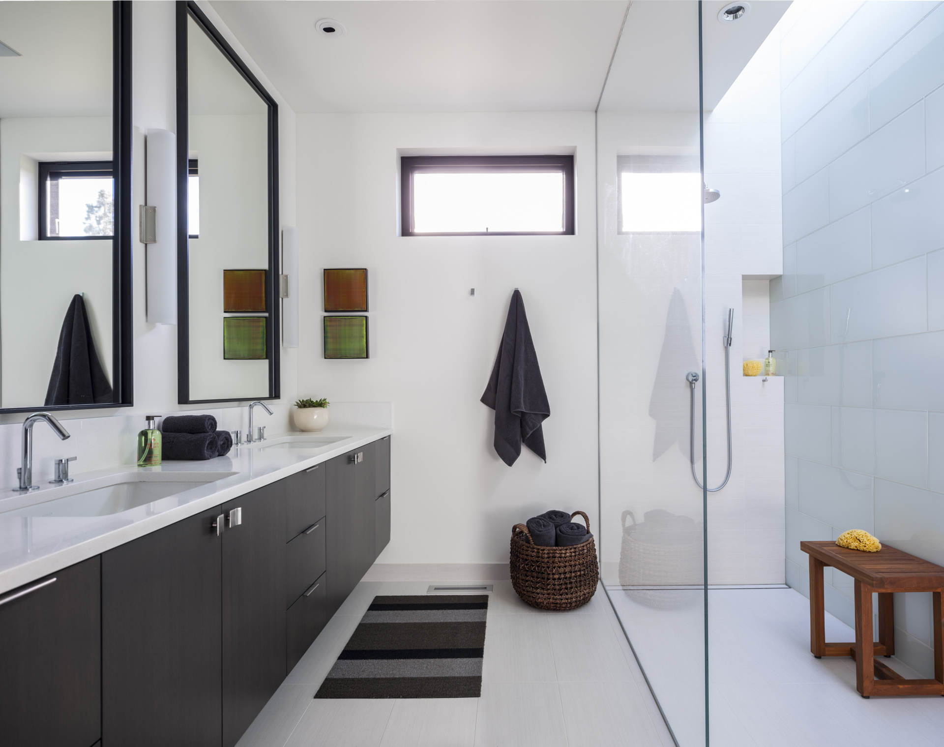 In this bathroom, crisp white walls and oversized tile are paired with a deep espresso wood vanity, while brushed chrome fixtures, a striped rug, and a woven basket add texture.