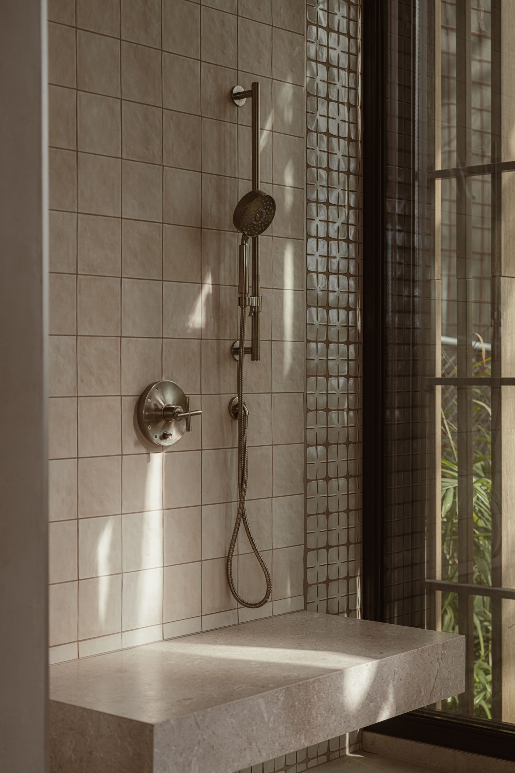 The ensuite continues the tonal calm, blending creamy handmade tiles with tactile stone finishes. A floating stone bench adds sculptural function to the open shower, where dappled light filters through the window.