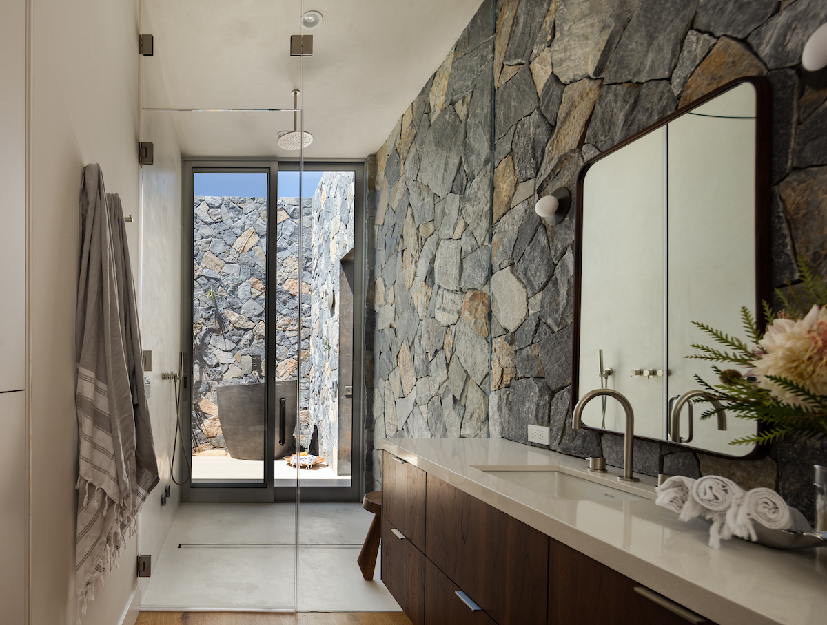 In this bathroom, the interior stone wall transitions to outside, where a deep soaking tub can be found. This private outdoor area is accessed via a glass door in the shower.