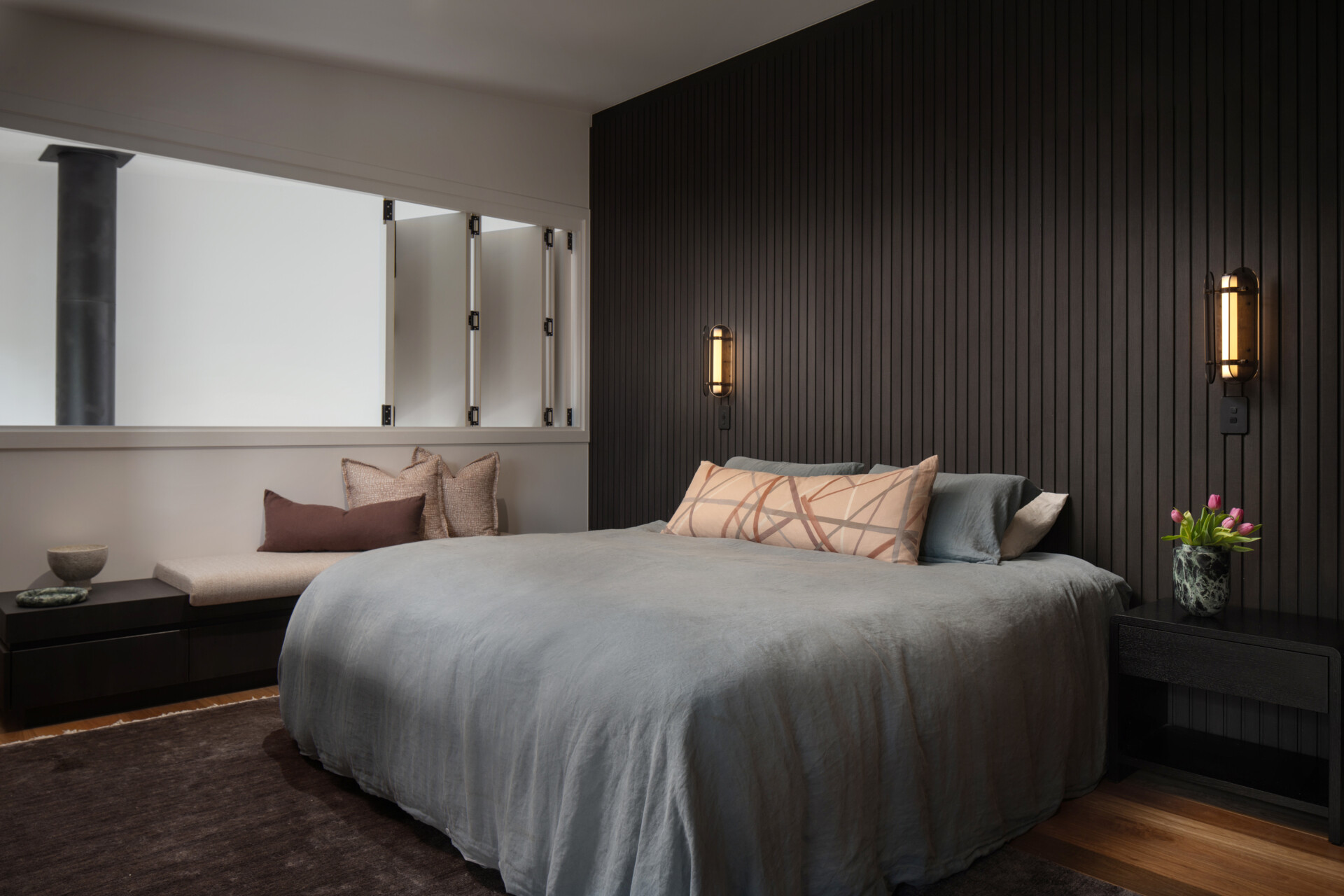 The bedroom balances modern sophistication with a warm, restful atmosphere. A dark vertical timber-panelled feature wall adds depth, softened by the glow of brass wall lights. A bench with textured cushions creates a comfortable reading nook, while fresh tulips on the bedside table bring a gentle touch of colour to the grounded palette.
