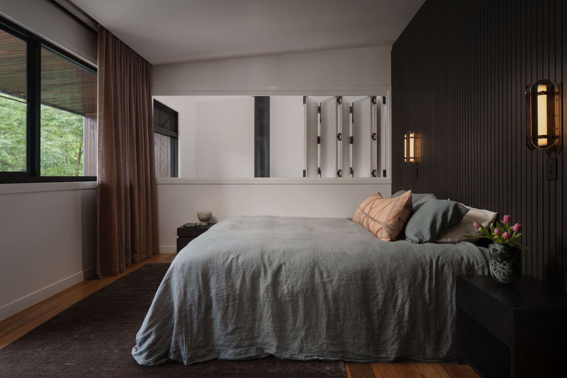 The bedroom balances modern sophistication with a warm, restful atmosphere. A dark vertical timber-panelled feature wall adds depth, softened by the glow of brass wall lights. A bench with textured cushions creates a comfortable reading nook, while fresh tulips on the bedside table bring a gentle touch of colour to the grounded palette.