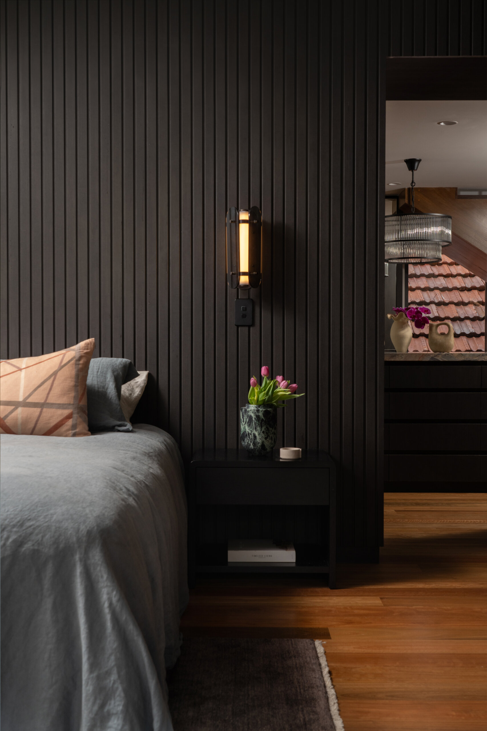 The bedroom balances modern sophistication with a warm, restful atmosphere. A dark vertical timber-panelled feature wall adds depth, softened by the glow of brass wall lights. A bench with textured cushions creates a comfortable reading nook, while fresh tulips on the bedside table bring a gentle touch of colour to the grounded palette.