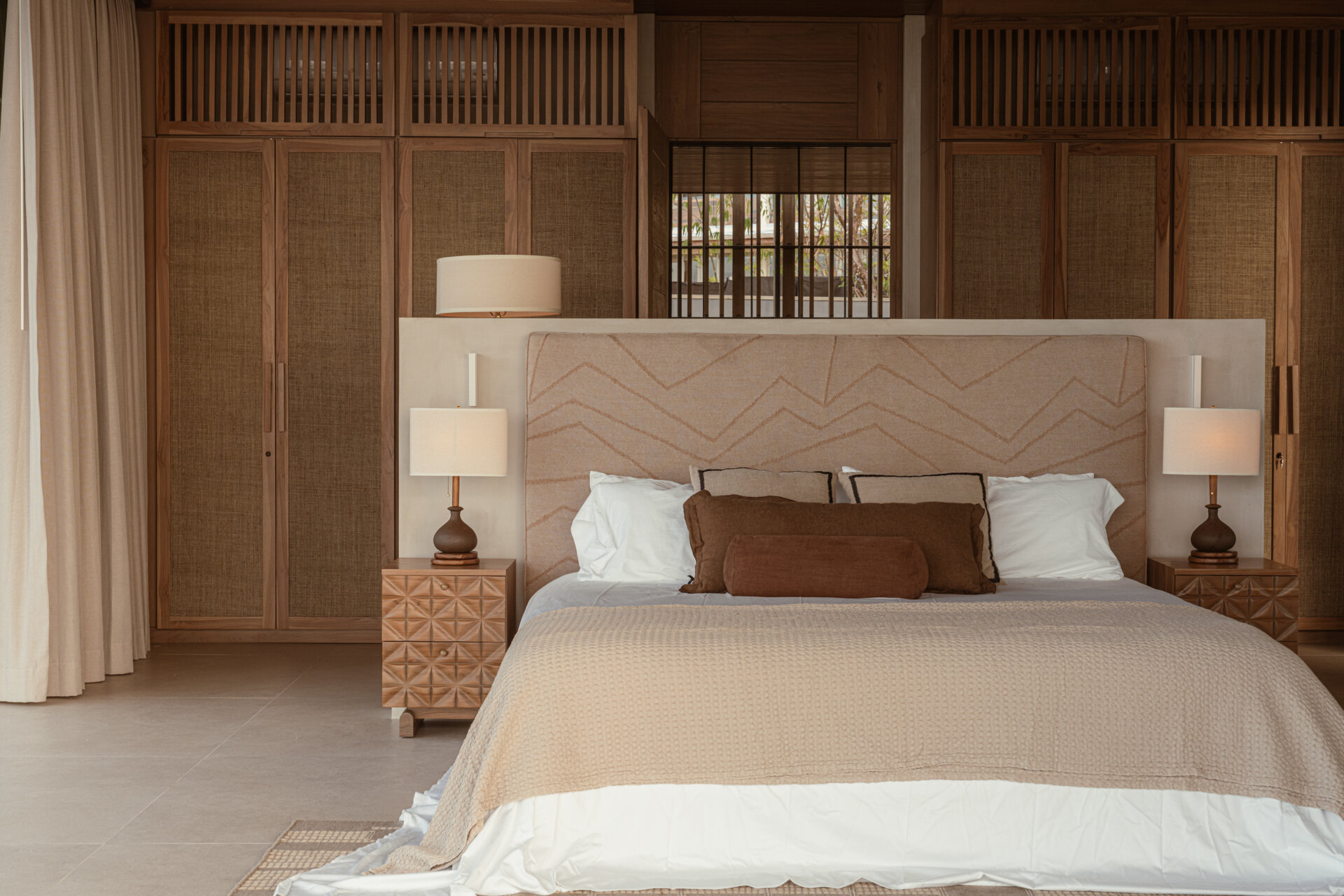Warm earth tones and layered textures give this bedroom and its ensuite a peaceful quality. The bedroom features a soft beige headboard with subtle geometric stitching, paired with woven wood panels and detailed nightstands that add a handcrafted touch. 