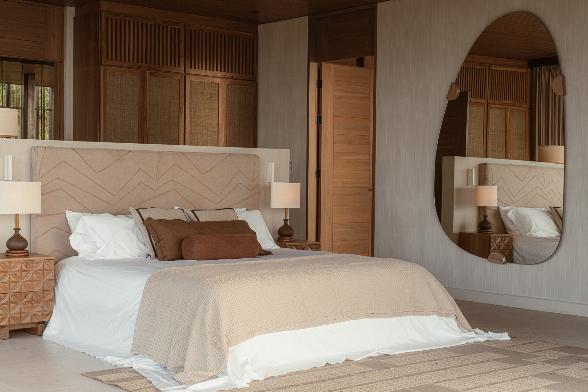 Warm earth tones and layered textures give this bedroom and its ensuite a peaceful quality. The bedroom features a soft beige headboard with subtle geometric stitching, paired with woven wood panels and detailed nightstands that add a handcrafted touch. 