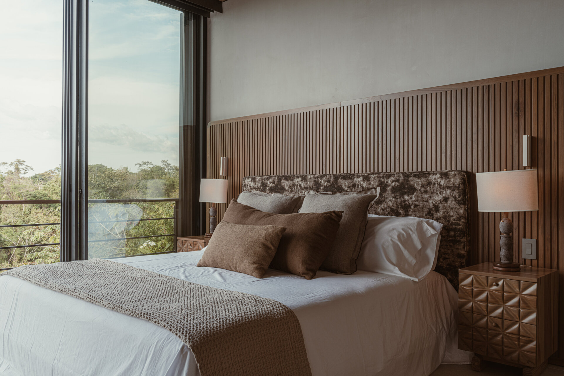 Natural materials lead the experience in this bedroom: reforested teak clads the ceiling and feature wall, while earthy stone flooring and soft woven textures ground the space. 