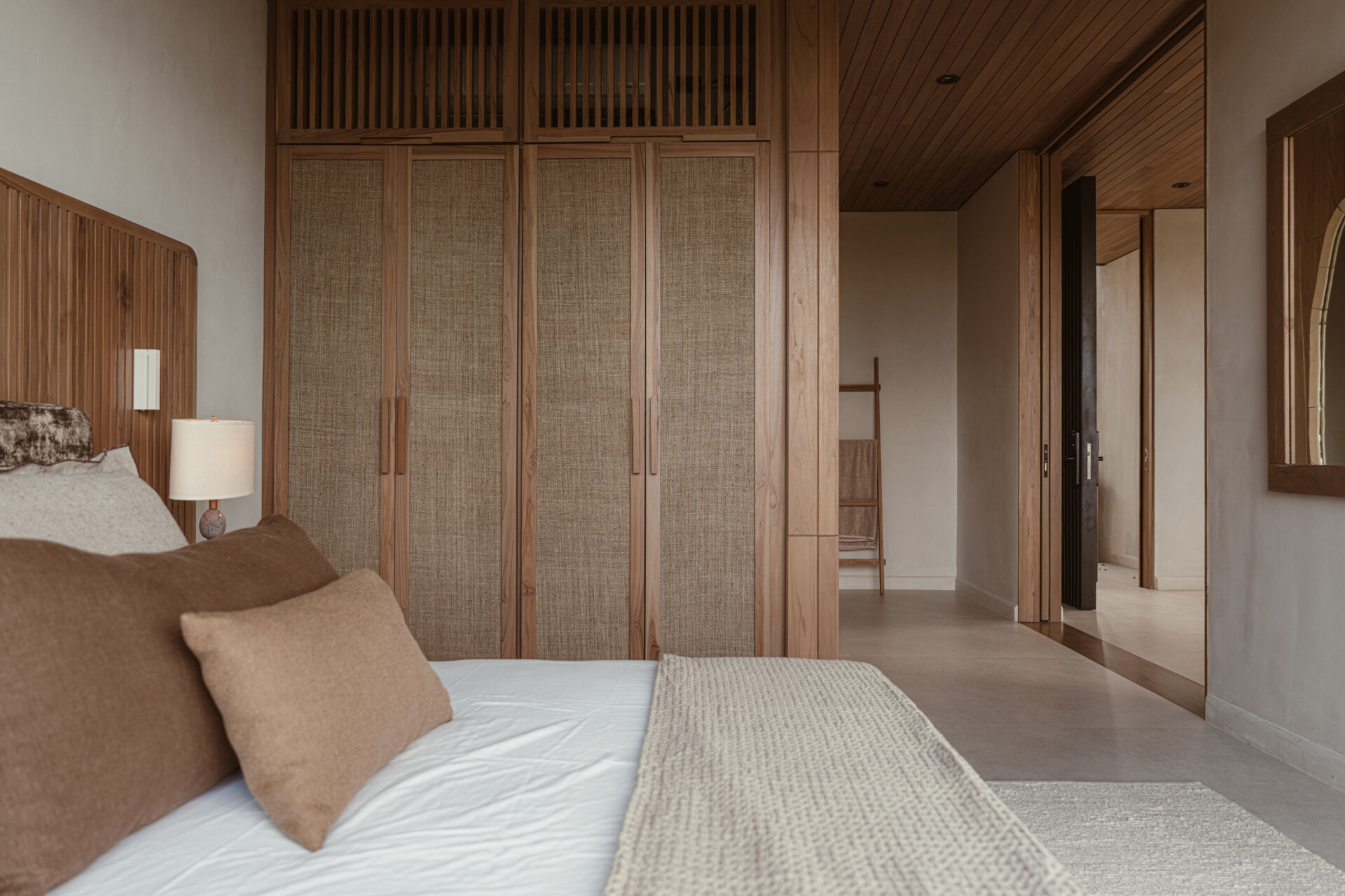 Natural materials lead the experience in this bedroom: reforested teak clads the ceiling and feature wall, while earthy stone flooring and soft woven textures ground the space. 