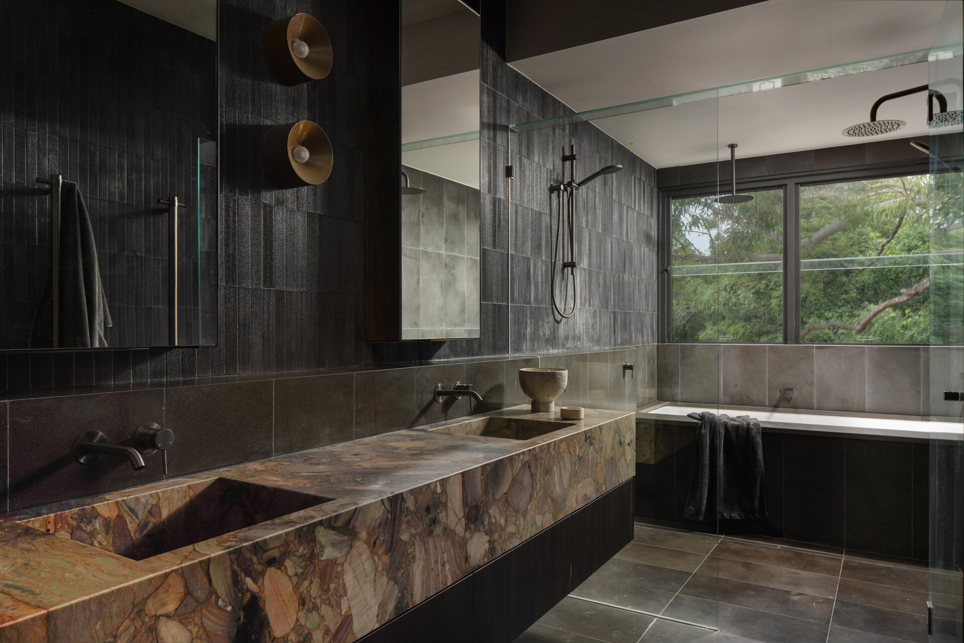 This bathroom pairs natural textures with clean, modern lines to create a serene retreat. Deep charcoal tiles wrap the walls and floor, their muted sheen reflecting daylight from a wide window that frames the surrounding greenery. 