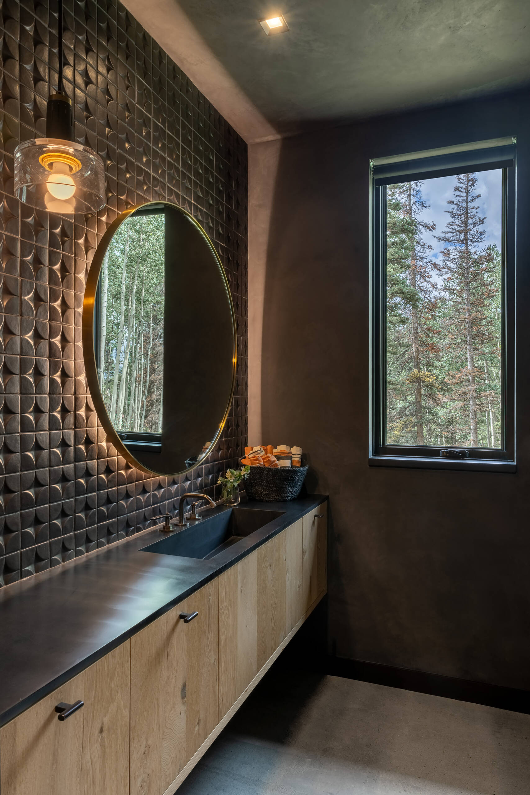 This bathroom pairs bold design with a sense of calm. Matte black walls and a sculpted tile backsplash create rich texture, while the round backlit mirror adds a soft glow that balances the moody palette. Warm wood cabinetry brings a natural touch, and the sleek countertop keeps the look streamlined.