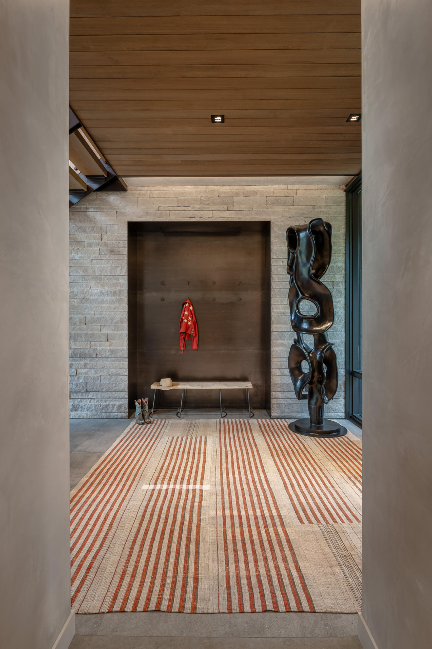 This entryway sets the tone with a mix of raw texture and curated detail. A recessed nook features simple hooks for coats, anchored by a bench that brings warmth and function, while a bold sculptural piece stands nearby as a striking counterpoint to the clean limestone walls.