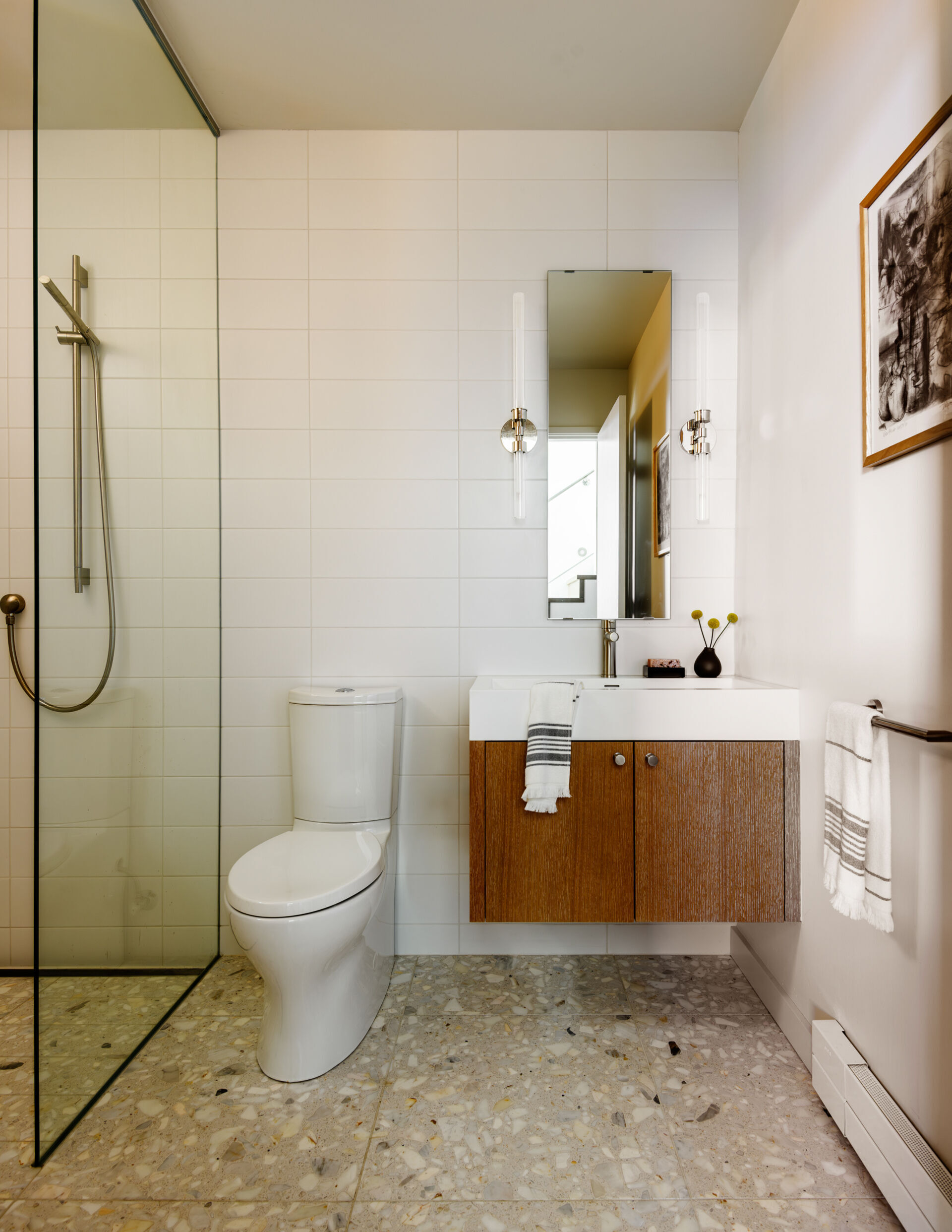 This bathroom features clean, modern finishes with terrazzo flooring, tiled wall, and a floating vanity. A slim mirror with vertical sconces enhances the sense of height, while simple details keep the space bright, efficient, and functional.