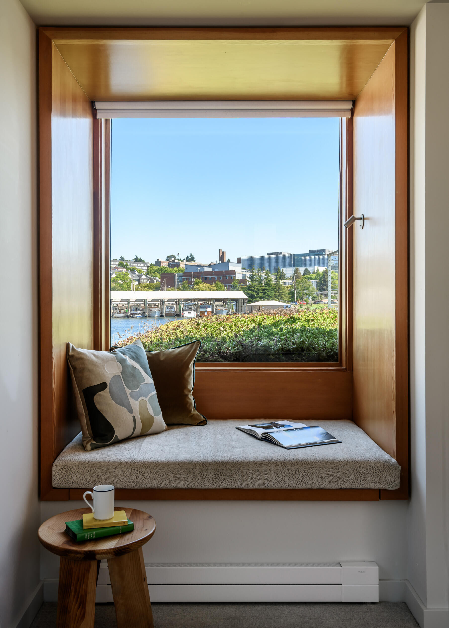 This primary bedroom in a float home offers more enclosure and intimacy. A wood headboard lines the wall and compliments the wood details found on the lower level, while a window seat overlooks the green roof, giving the bedroom a quiet view softened by planted greenery.
