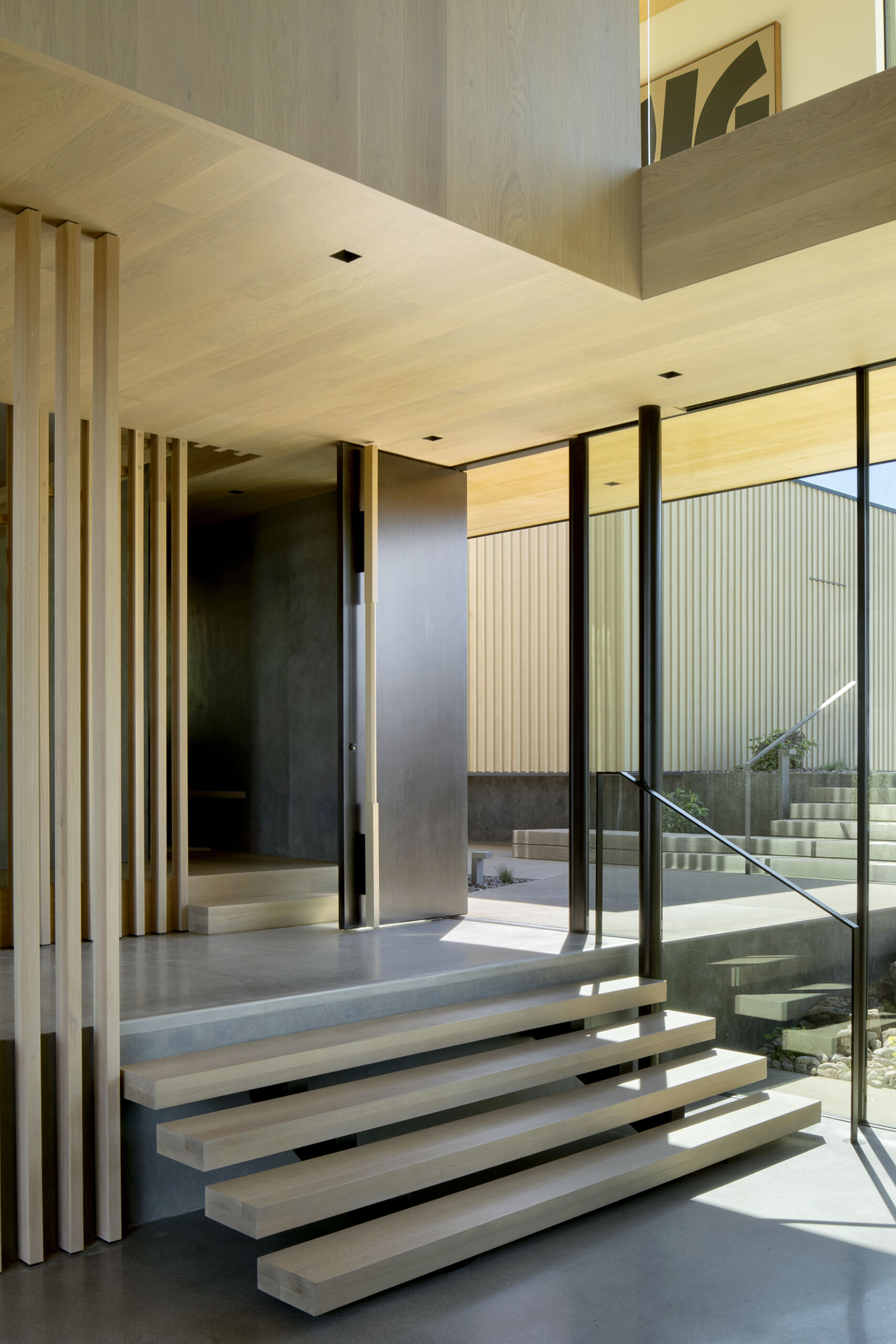 A large patinated steel pivot door welcomes visitors beneath the overhang, opening into an intermediate level with framed eastern views and a tactile connection to the outdoors.