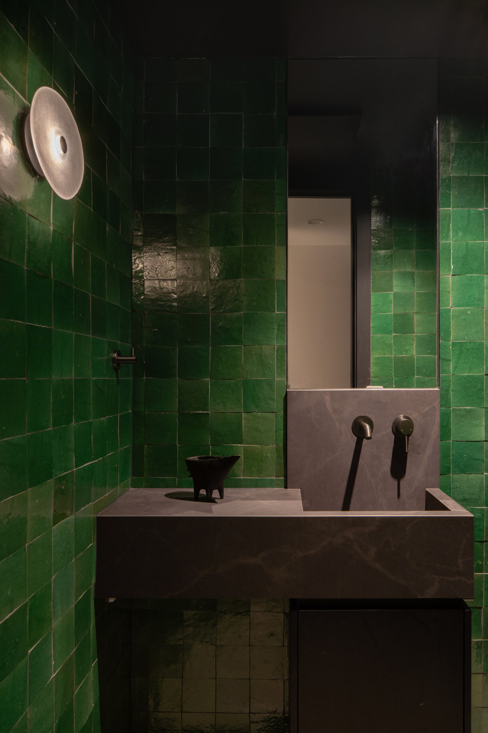 Glossy deep emerald tiles wrap the walls in this bathroom, their surface catching the light to create gentle shifts in tone. A charcoal stone vanity introduces clean lines and a sense of refinement, complemented by matte black fixtures. The combination of rich color and smooth, modern finishes gives the space a moody, intimate character with a quietly dramatic edge.