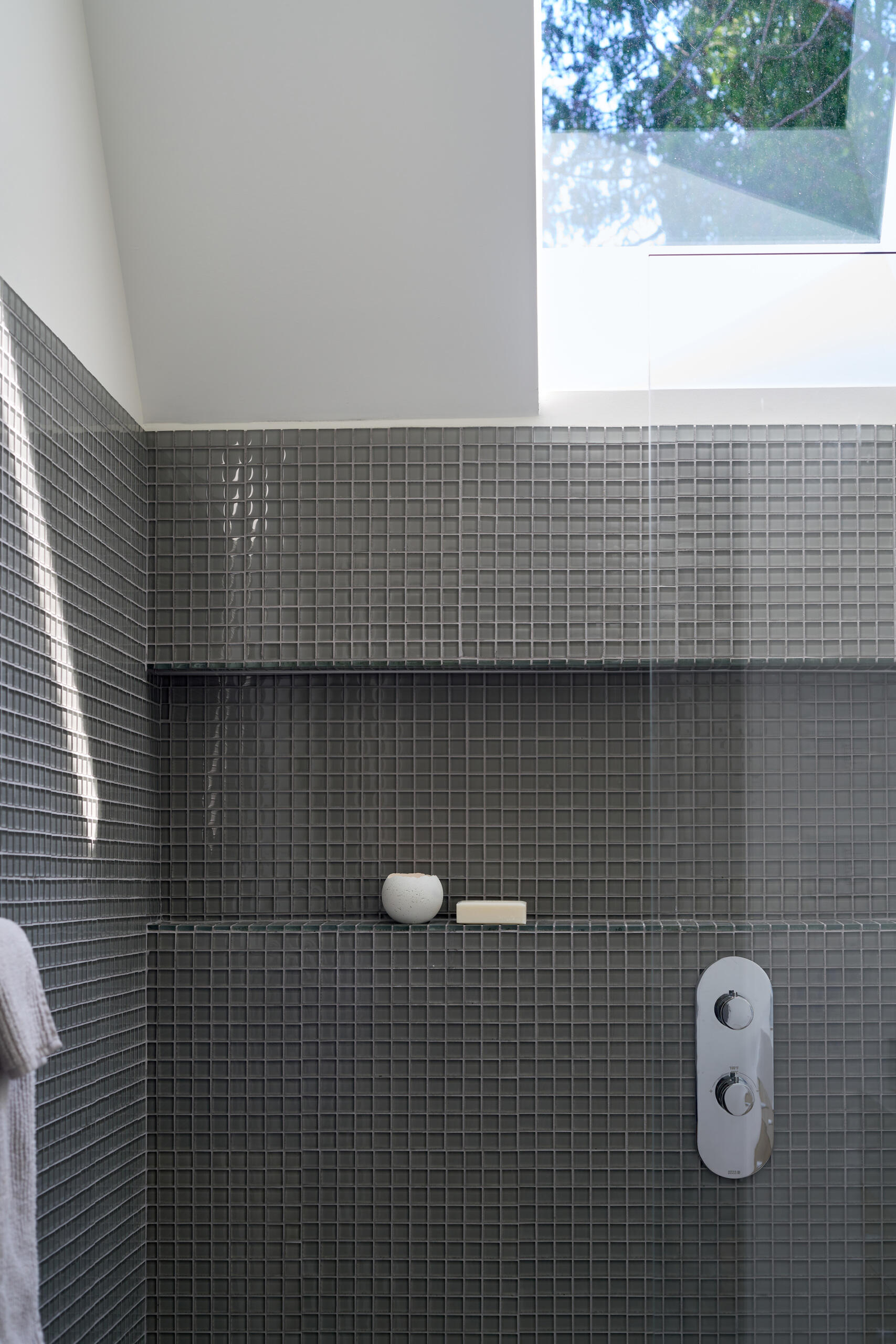 In this modern bathroom, small grey tiles line the shower and shelving niche, while a skylight adds natural light.