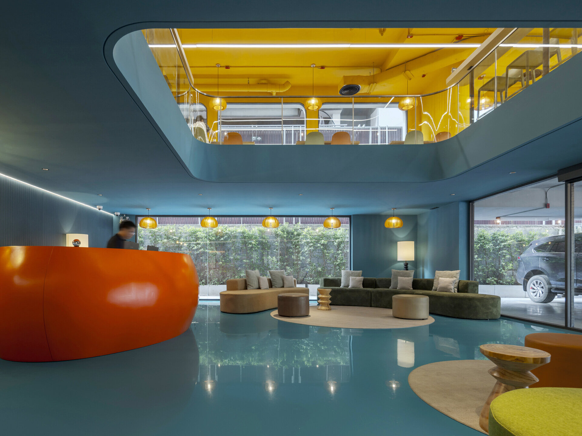 Step into a bold and playful lobby where a sculptural orange reception desk pops against lush colorful walls, all beneath soaring ceilings and layered views of the common areas above.