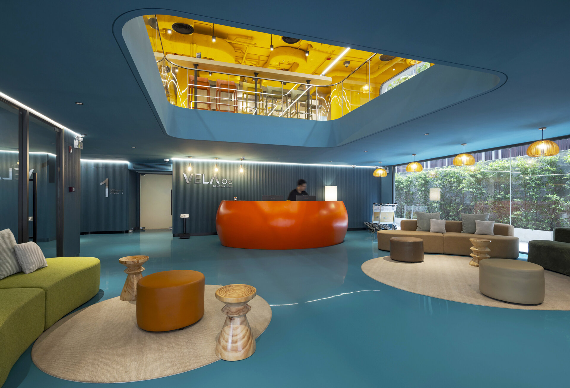 Step into a bold and playful lobby where a sculptural orange reception desk pops against lush colorful walls, all beneath soaring ceilings and layered views of the common areas above.