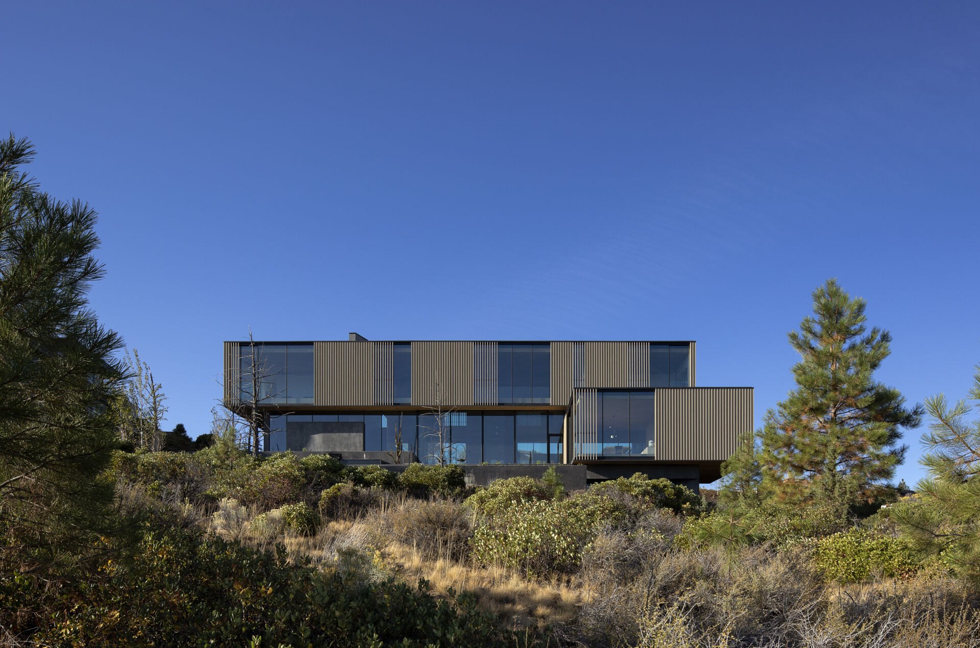 This modern home is designed as a series of cascading platforms that step down a pumice-covered hillside. Inspired by nearby volcanic rock formations, faceted mass walls anchor the structure, guiding movement and framing both intimate and expansive views. 