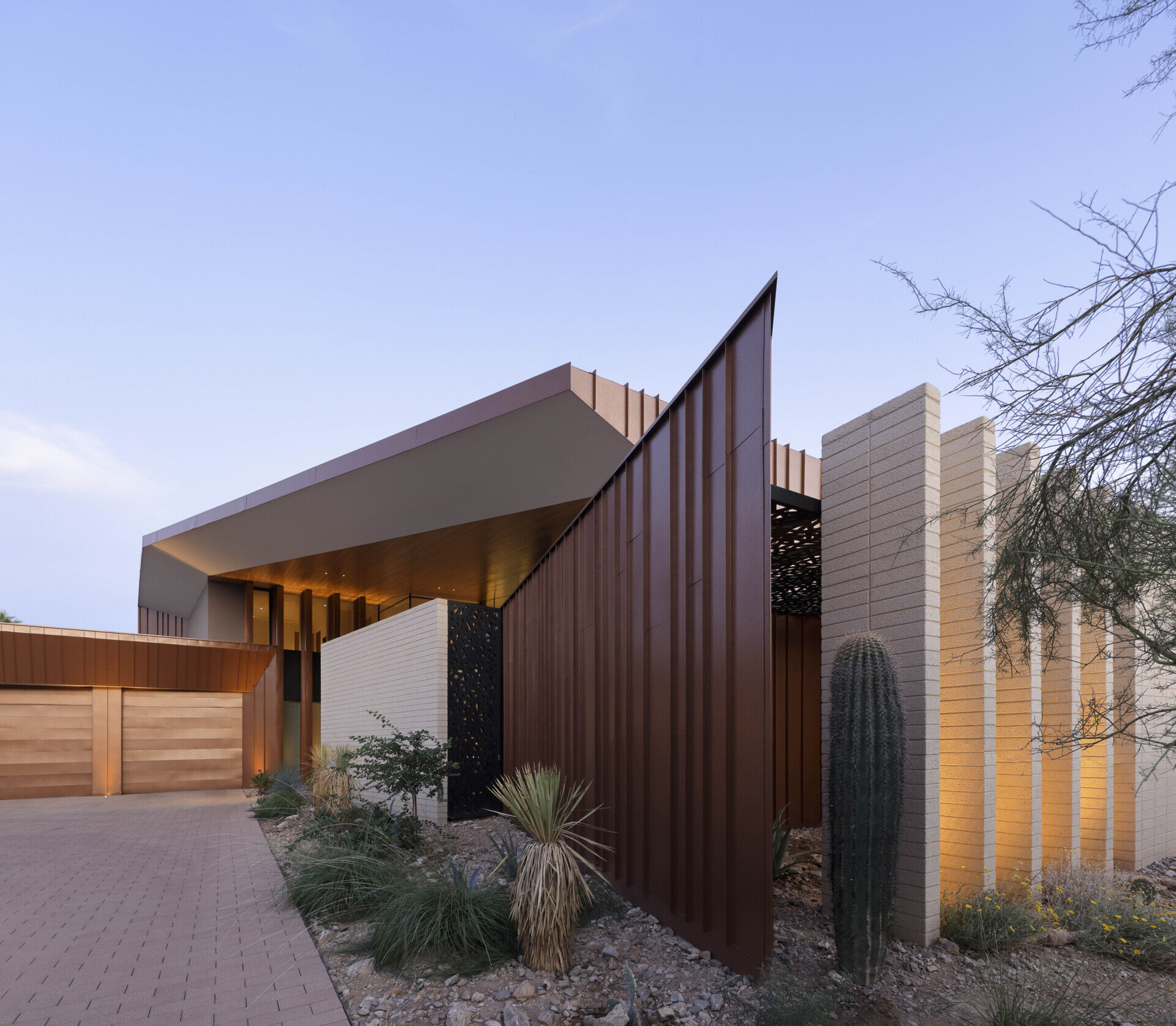 Defined by folding metal planes and rugged blasted-face masonry, the architecture of this modern house recalls the layered beauty of a geode. The weathered metal acts as a protective shell, sheltering the glass-filled interior that glows with natural daylight.