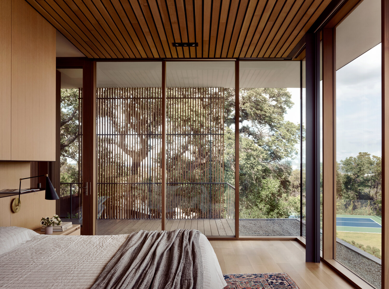 This bedroom features floor-to-ceiling windows with wood slats that filter light, pairing warm timber finishes with soft textures for a serene connection to the outdoors.