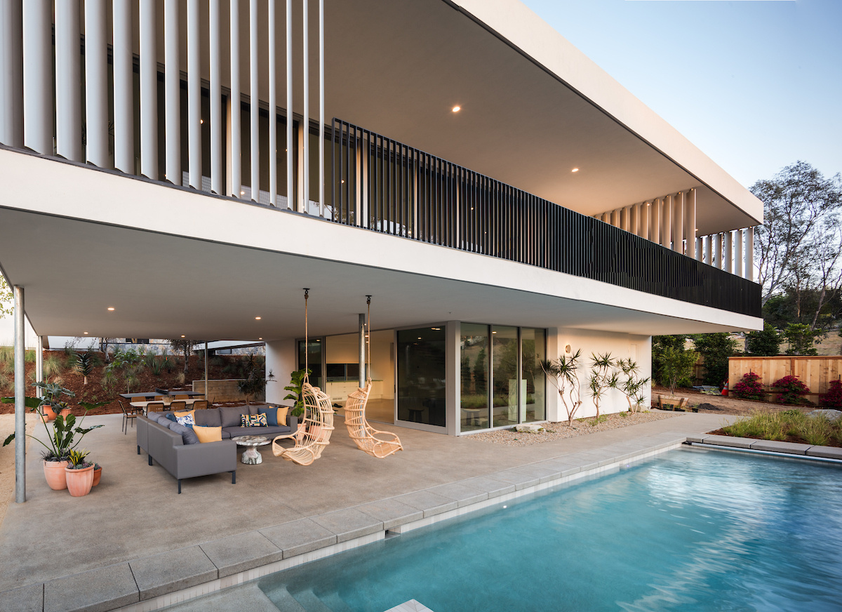 A modern house with a white exterior has a covered outdoor living room by a pool.