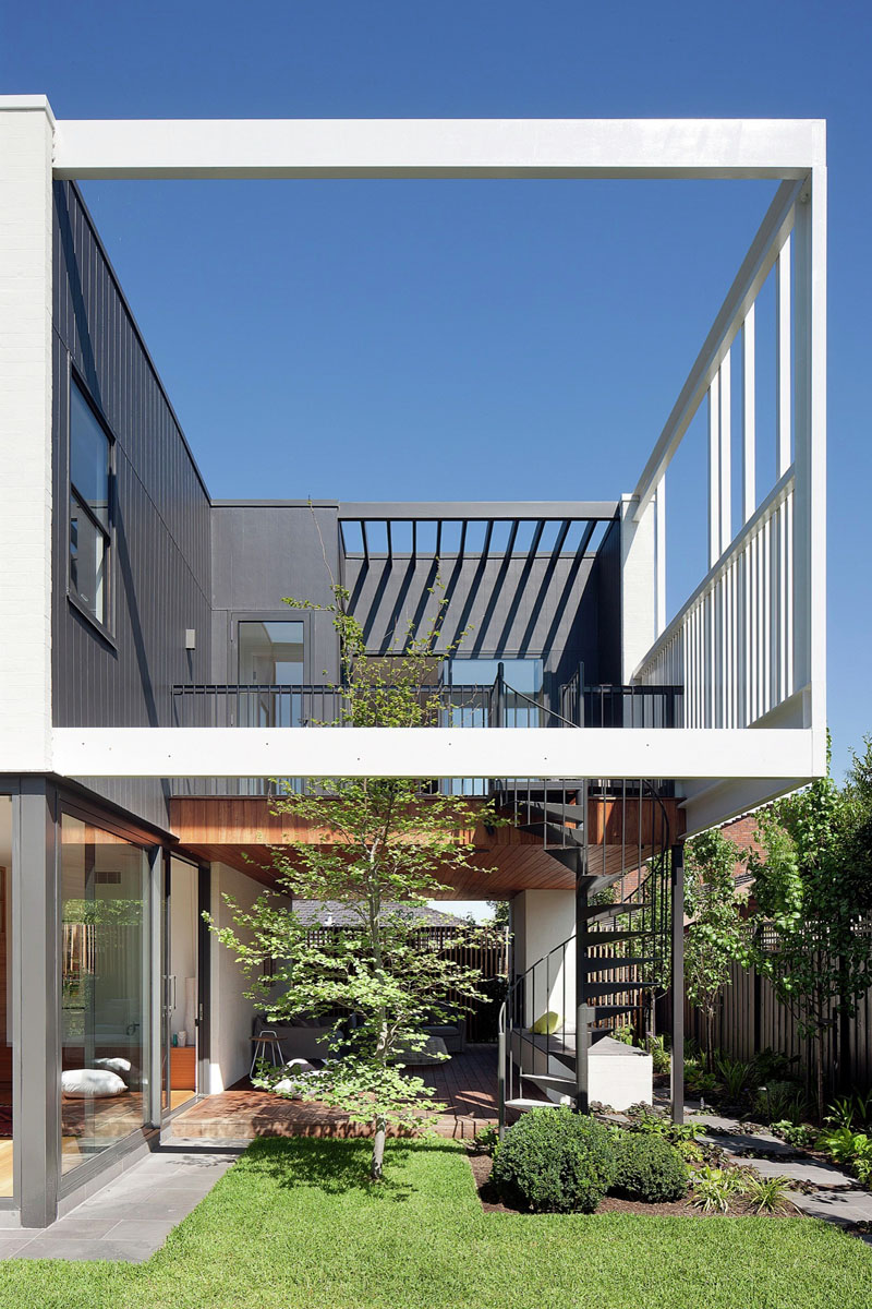 A modern home with a covered outdoor lounge framed by greenery, complete with a built-in fireplace and wood storage.