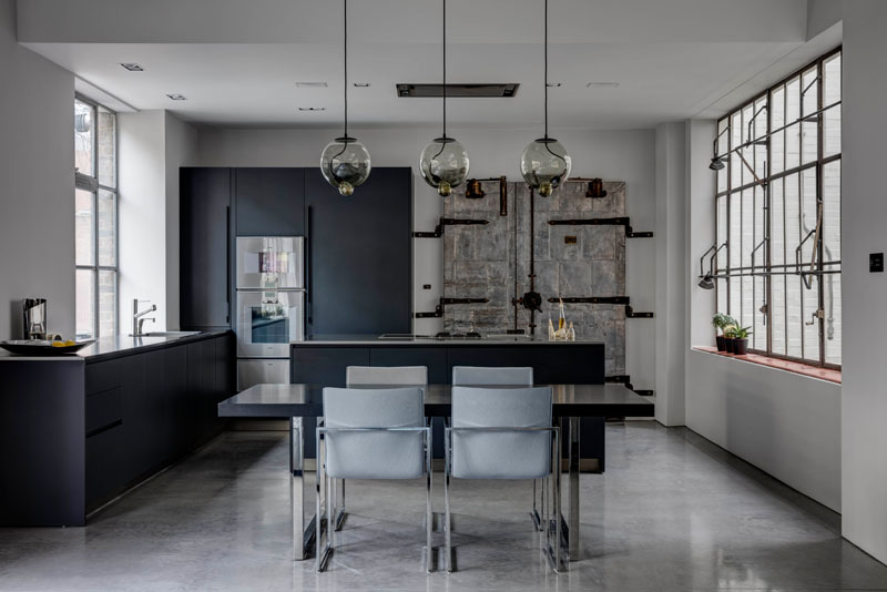 In the kitchen, a striking old industrial door has been incorporated into the design, tying the home back to its factory roots. Black cabinetry contrasts against the crisp white walls and polished concrete flooring, while the warm tones of wood and subtle industrial hues add depth.