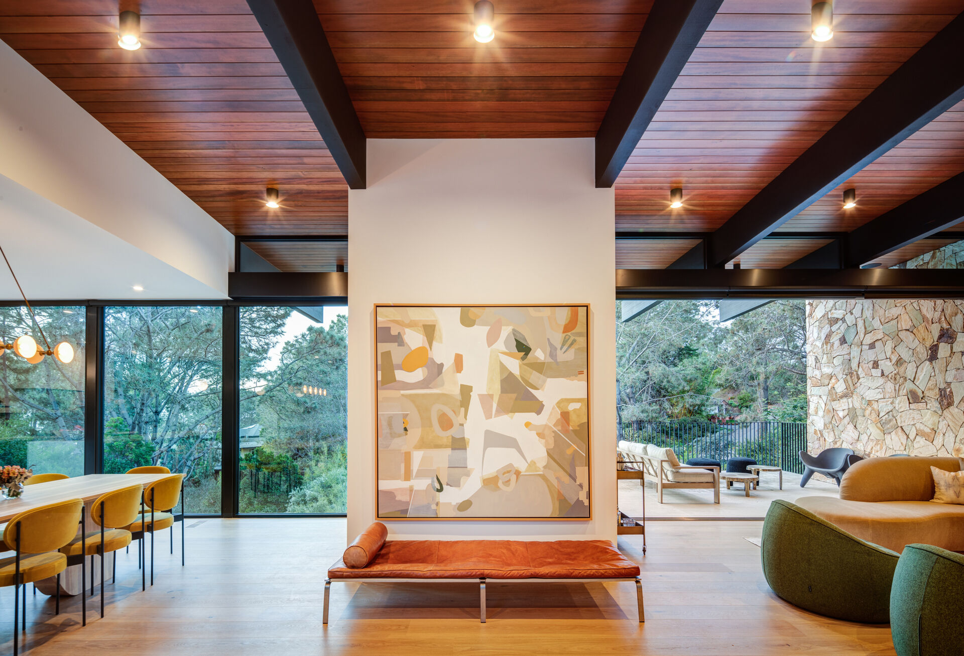 At the top of the stairs, a rust-colored bench and artwork anchors the social areas of the home, tying together warm wood ceilings, stone accents, and earthy tones that echo the landscape beyond the glass walls.