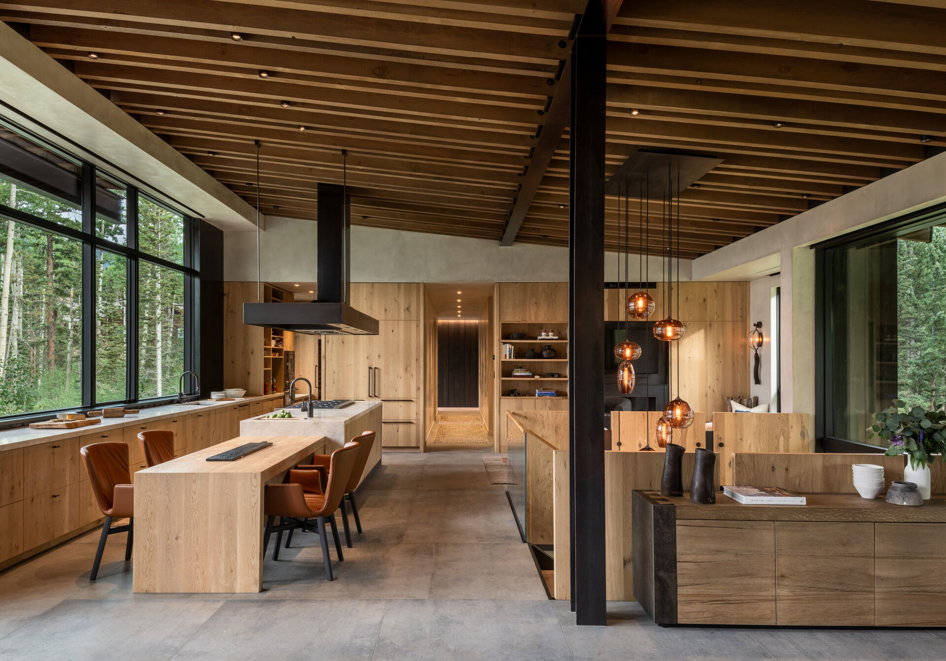 This kitchen centers around an island with a stove, range, and sink. Integrated storage and a refrigerator clad in vertical grain oak veneer with Emtek hardware continue the theme of craftsmanship. A custom hood vent hovers as a focal point above the island, while the open floor plan ensures the dining and living spaces flow together seamlessly.