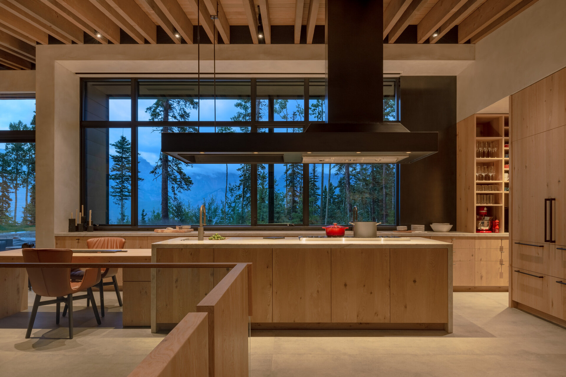 This kitchen centers around an island with a stove, range, and sink. Integrated storage and a refrigerator clad in vertical grain oak veneer with Emtek hardware continue the theme of craftsmanship. A custom hood vent hovers as a focal point above the island, while the open floor plan ensures the dining and living spaces flow together seamlessly.