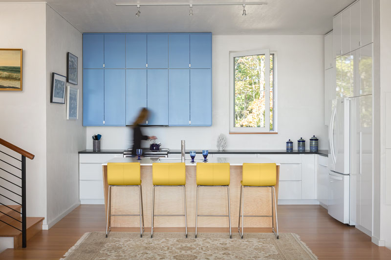 The kitchen brings a lively contrast to the home’s calm interiors. Bright yellow bar stools energize the island, while sky-blue cabinetry adds both storage and a cheerful focal point. Clean white cabinets and counters keep the look fresh, and a window frames views of the trees outside.