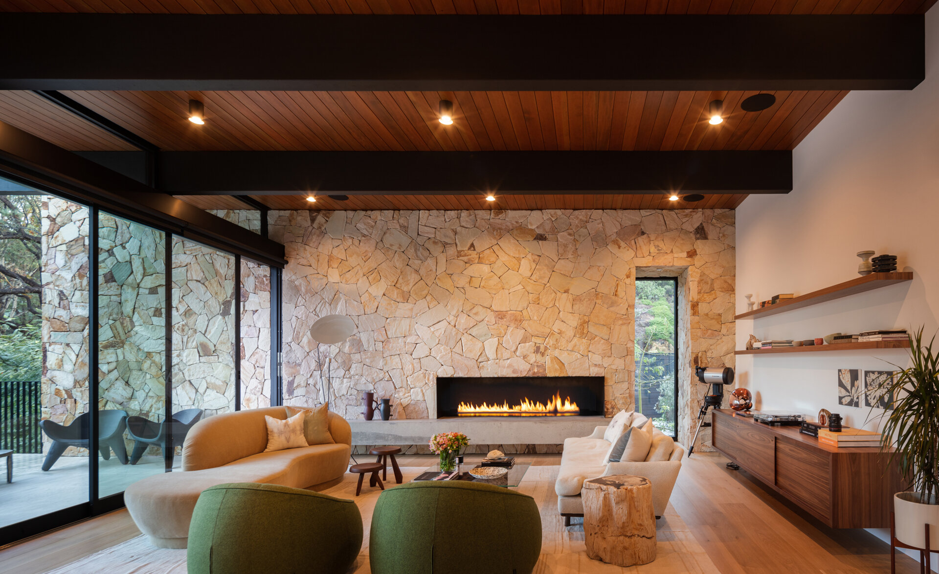 The living room centers around a stone wall and fireplace, while floor-to-ceiling glass walls dissolve the boundary between indoors and outdoors. Just beyond, a partially covered outdoor living room extends the gathering space into nature, perfect for both quiet evenings and entertaining.