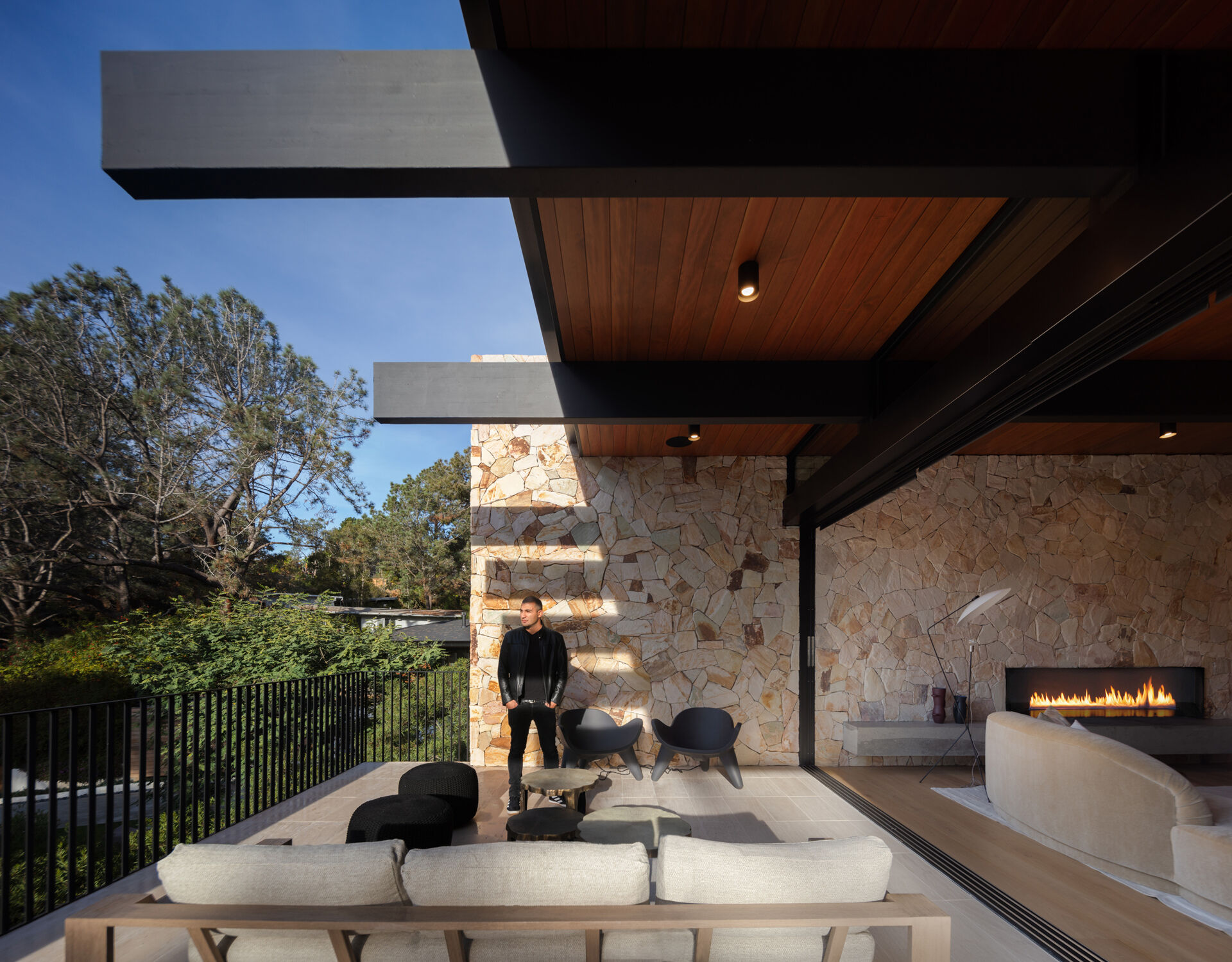 The living room centers around a stone wall and fireplace, while floor-to-ceiling glass walls dissolve the boundary between indoors and outdoors. Just beyond, a partially covered outdoor living room extends the gathering space into nature, perfect for both quiet evenings and entertaining.