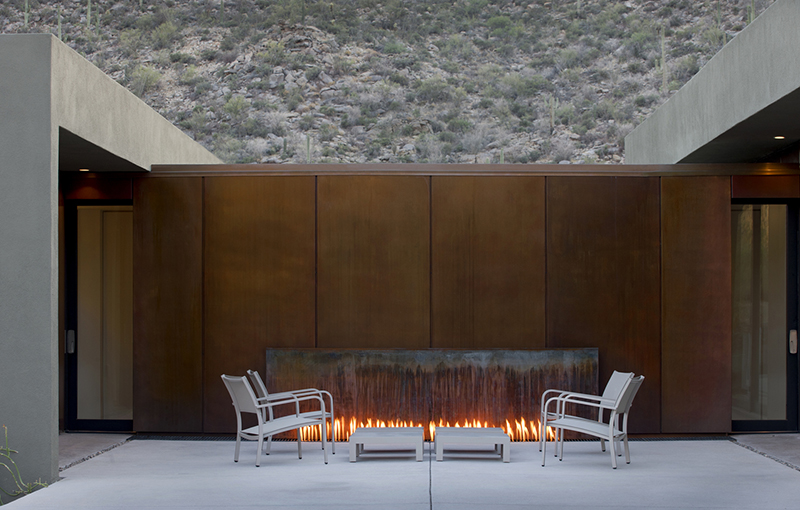 Between different areas of the house lies an outdoor fireplace framed by weathered steel. This gathering spot connects the pool and living spaces, adding warmth and character.