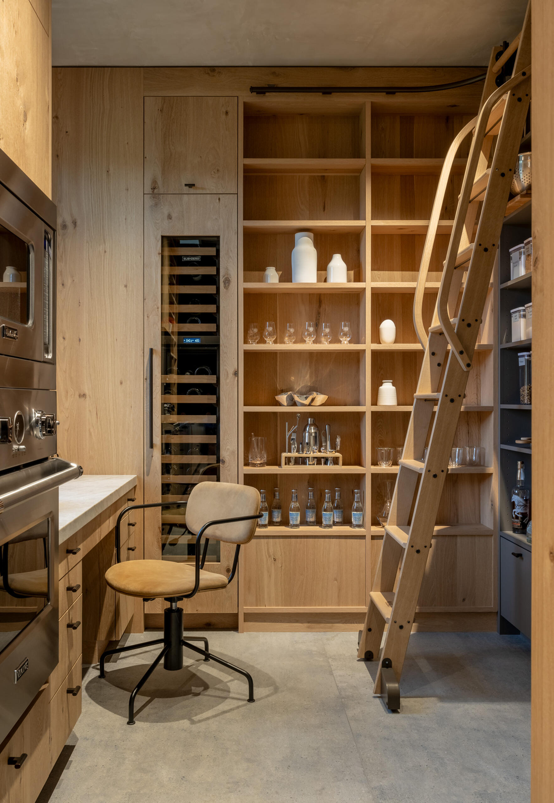This pantry, located off to the side of the kitchen, has full-height oak cabinetry surrounding the space, integrating open shelving for glassware and ceramics, while a built-in wine cooler adds a touch of sophistication, and a desk creates a workspace. A rolling ladder provides easy access to upper shelves, blending functionality with charm.