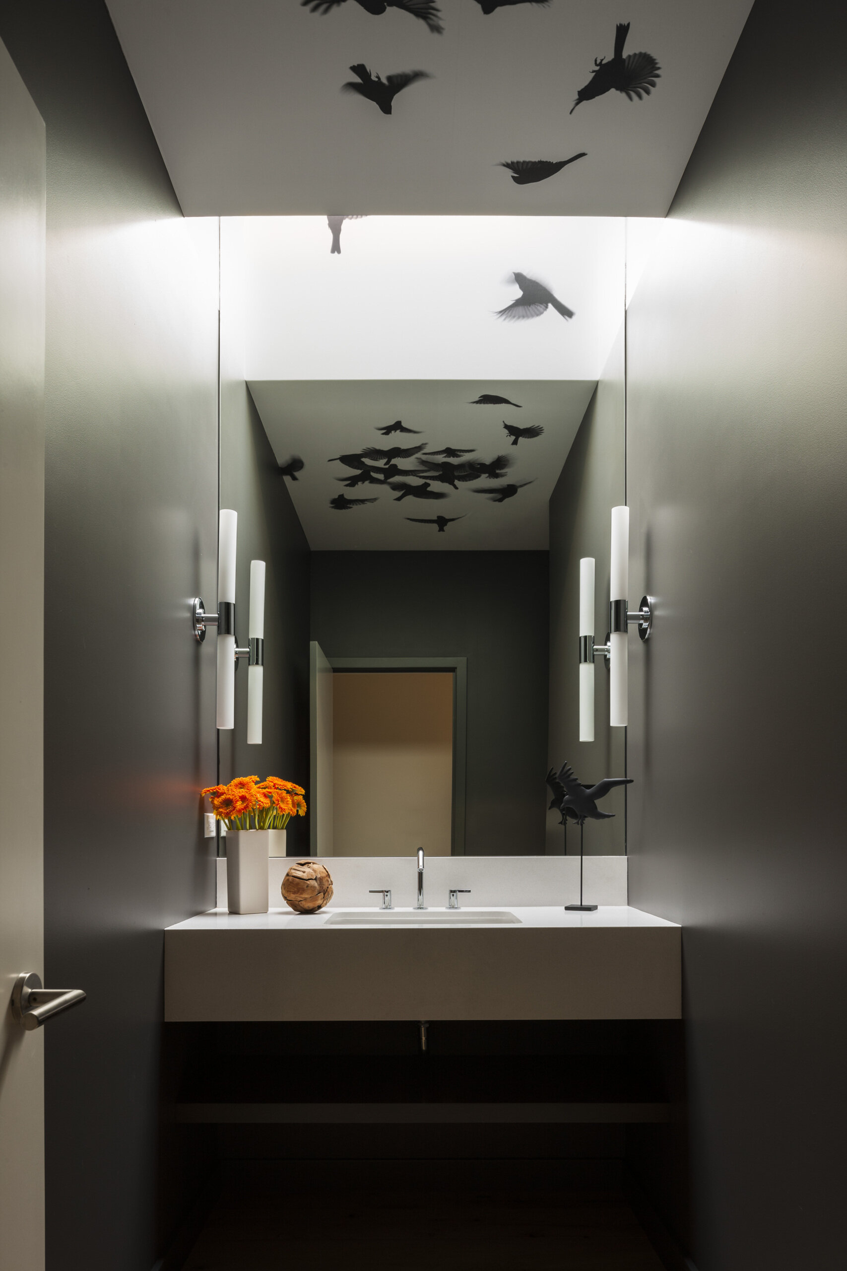 Moody charcoal walls set a quiet backdrop for this powder room, while a crisp floating white vanity keeps the space feeling open and easy to maintain. A luminous ceiling panel washes the room in soft light, revealing a flock of black birds that turns the ceiling into artwork.