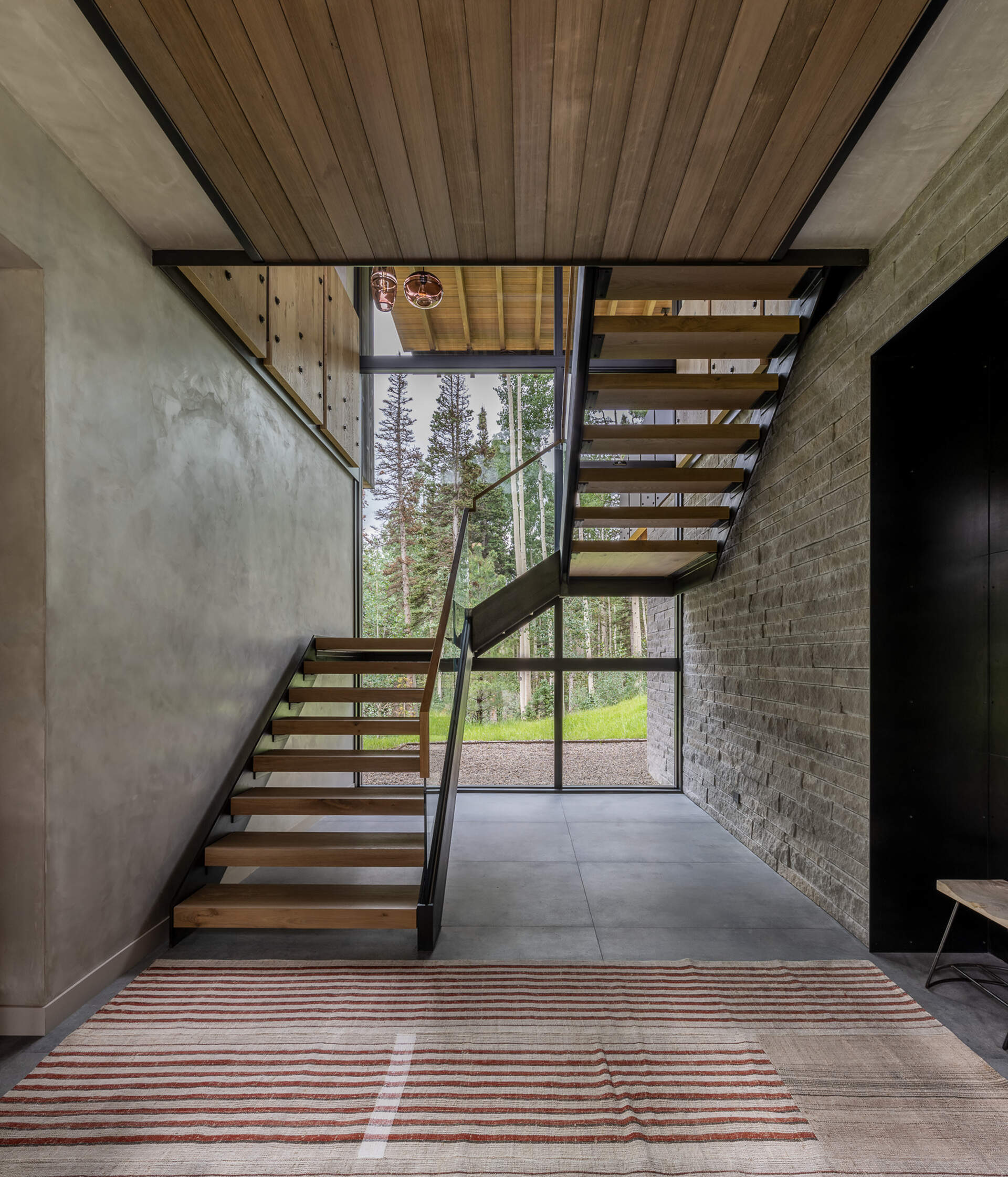 Just beyond the entry, a crafted staircase leads upward, designed as both functional and sculptural. It stands as a centerpiece that draws the eye toward the forest backdrop beyond the glass wall.