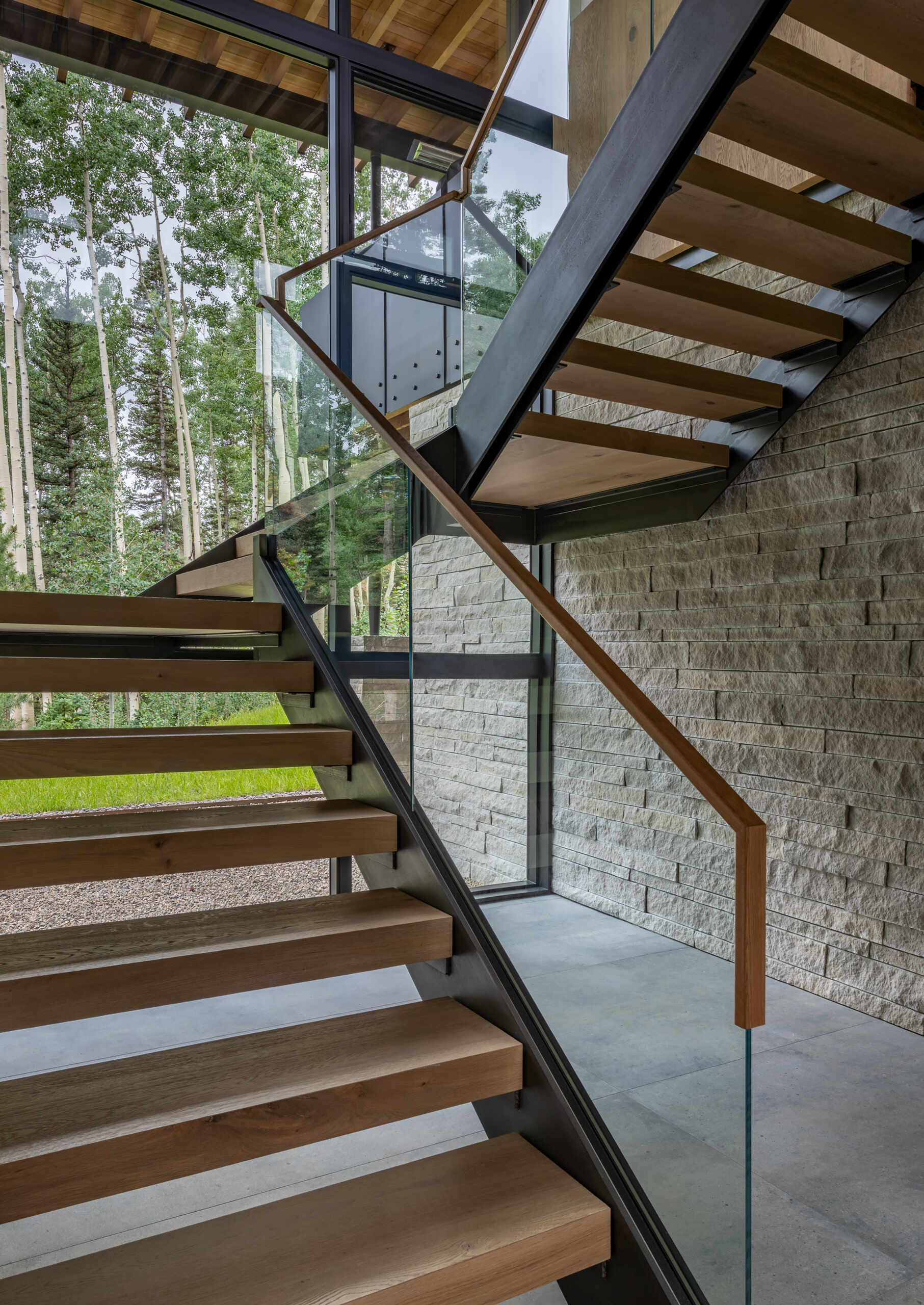 Just beyond the entry, a crafted staircase leads upward, designed as both functional and sculptural. It stands as a centerpiece that draws the eye toward the forest backdrop beyond the glass wall.