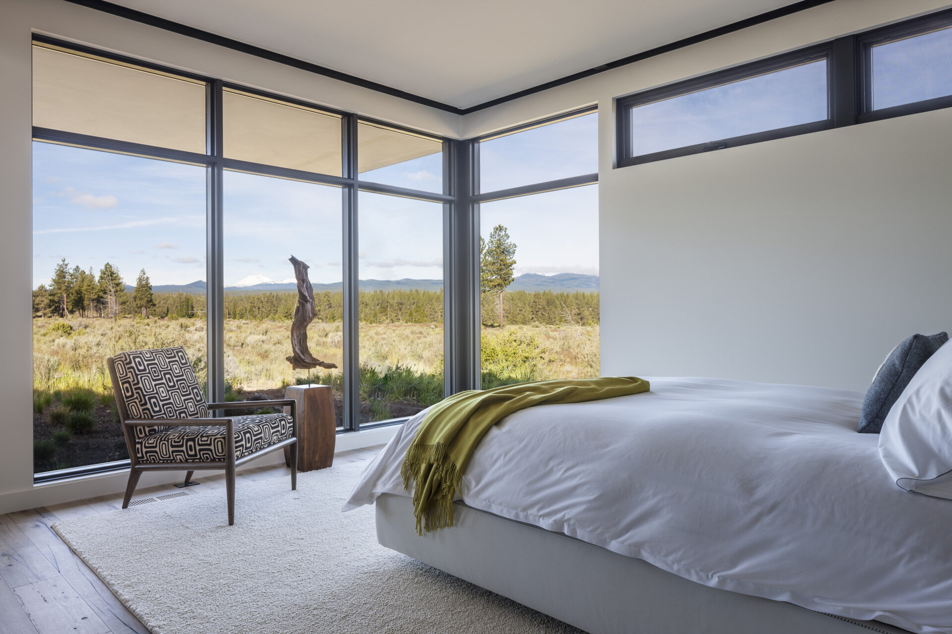 Wrapped in windows, this bedroom wakes to mountain views. Soft white walls and bedding keep the palette quiet, while a mossy green throw adds a relaxed hit of color and a mid-century chair with graphic upholstery creates character without clutter.