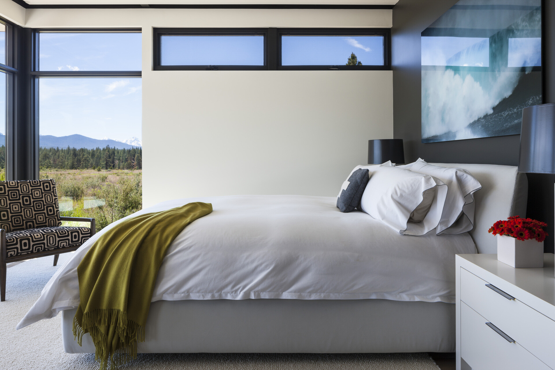 Wrapped in windows, this bedroom wakes to mountain views. Soft white walls and bedding keep the palette quiet, while a mossy green throw adds a relaxed hit of color and a mid-century chair with graphic upholstery creates character without clutter.
