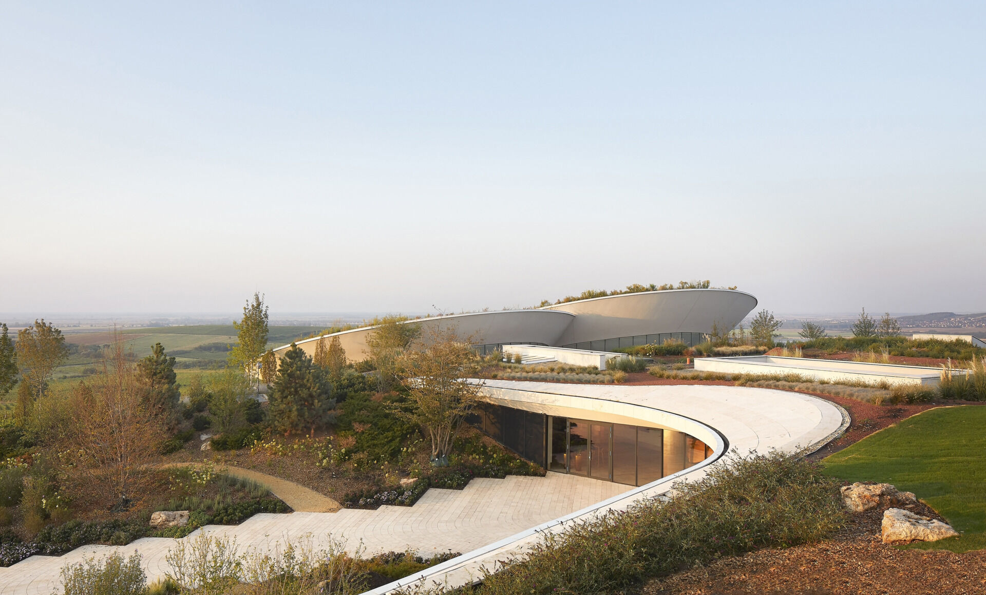 The winery’s lens-shaped floating volumes, covered in plants, create a striking landmark, balancing dramatic design with functional underground fermentation and storage areas.