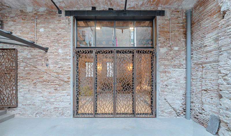 Original brick walls and a built-in sofa with colorful cushions leads to folding glass doors that open to a private courtyard, framed by decorative steel screens.