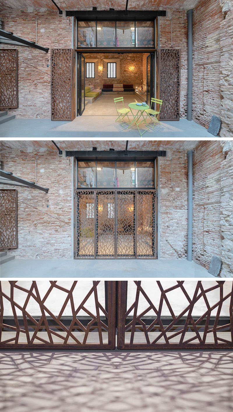 Original brick walls and a built-in sofa with colorful cushions leads to folding glass doors that open to a private courtyard, framed by decorative steel screens.