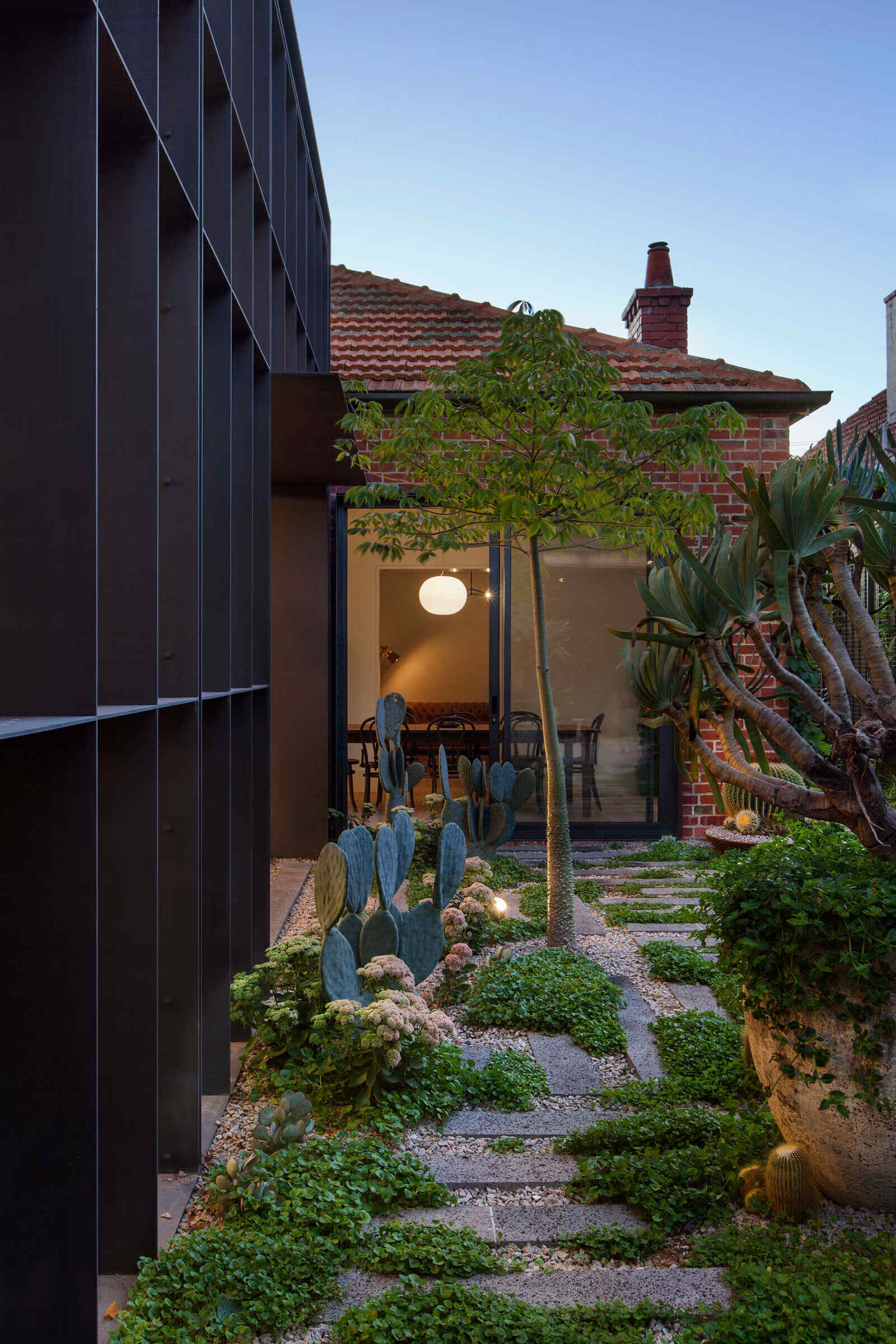 The path opens to a small pergola with a patio and seating, creating a peaceful outdoor threshold. The new pavilion’s structural steel baffle regulates sunlight while casting dramatic shadows.
