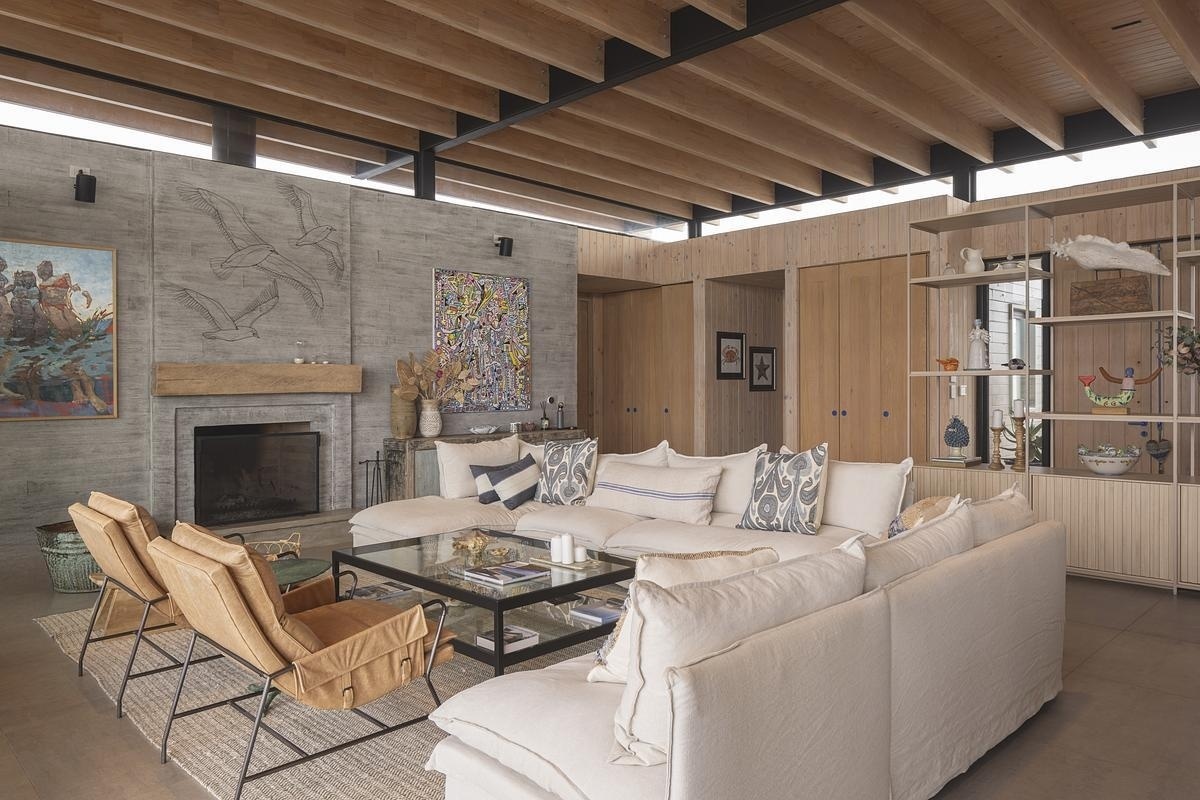 Living, dining, and kitchen flow together, each area defined yet connected. A fireplace anchors the living room, while a large wooden table marks the dining zone before the layout transitions seamlessly into the kitchen. Overhead, shifting light plays across the beams, guiding the gaze outward to the horizon.