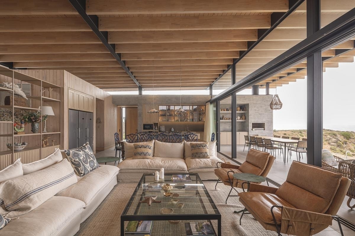 Living, dining, and kitchen flow together, each area defined yet connected. A fireplace anchors the living room, while a large wooden table marks the dining zone before the layout transitions seamlessly into the kitchen. Overhead, shifting light plays across the beams, guiding the gaze outward to the horizon.