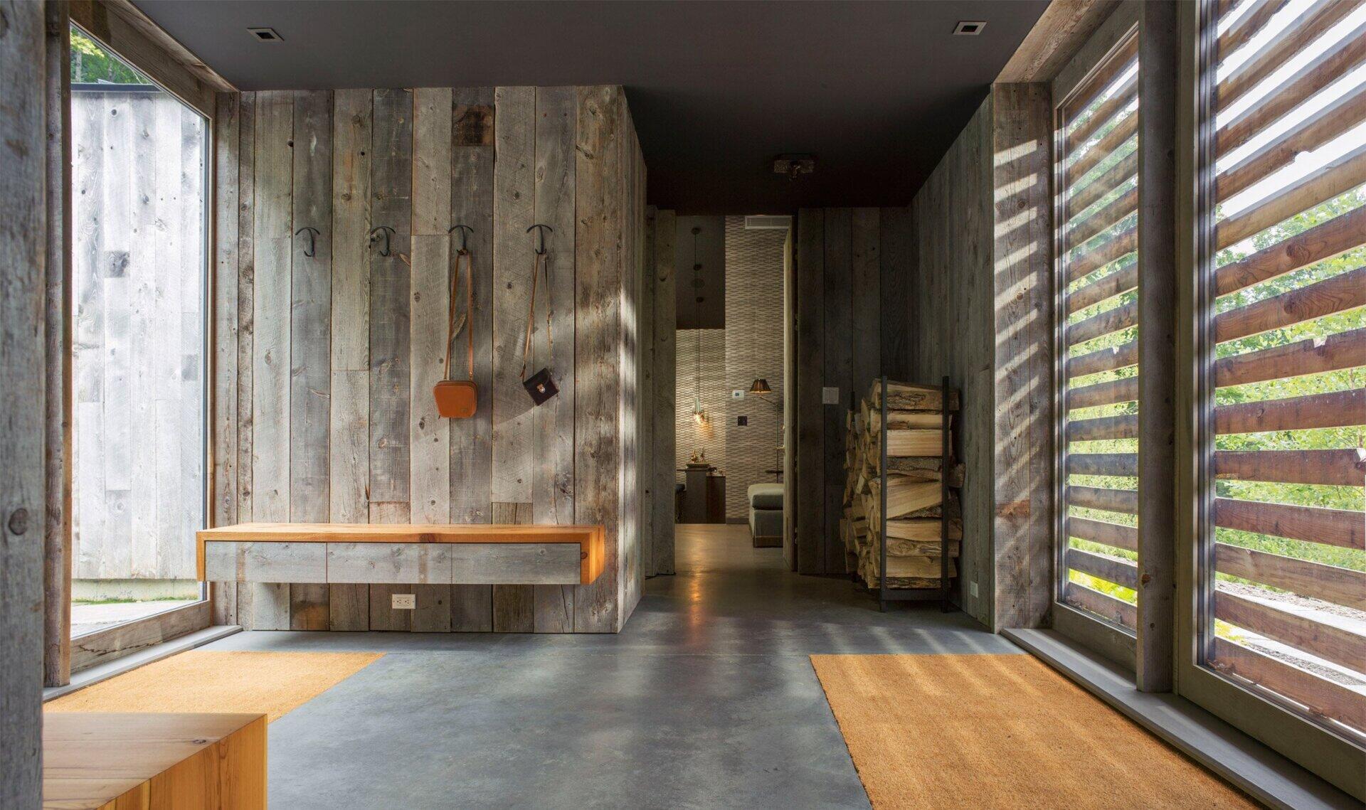 The siding doesn’t stop at the threshold. In the entryway, the boards continue seamlessly inside, wrapping the walls and even forming built-in benches. From here, a view opens directly through the house to the terrace and forest beyond.