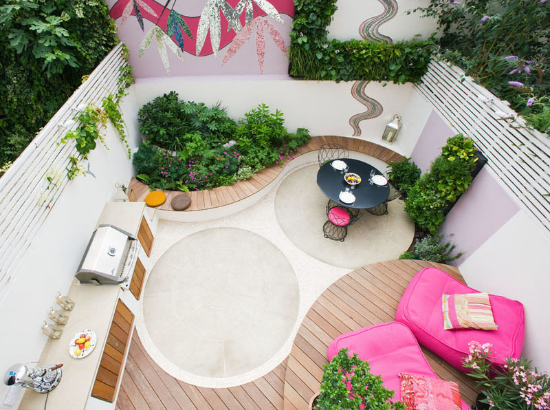An Ibiza-inspired garden makeover featuring a vibrant mosaic wall, bespoke outdoor kitchen with BBQ, York stone dining area, handcrafted hardwood bench, and a lush herb wall. Bright pink lounge chairs and evergreen planting create a colorful, family-friendly backyard perfect for entertaining.