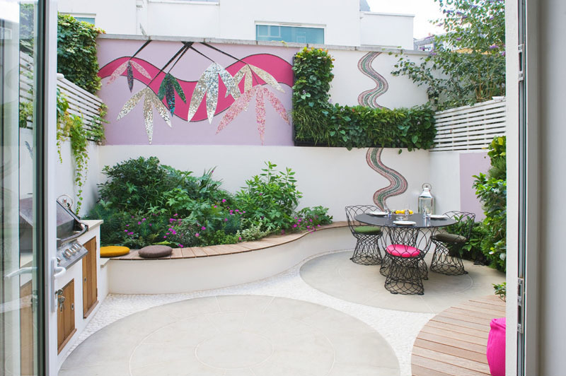 An Ibiza-inspired garden makeover featuring a vibrant mosaic wall, bespoke outdoor kitchen with BBQ, York stone dining area, handcrafted hardwood bench, and a lush herb wall. Bright pink lounge chairs and evergreen planting create a colorful, family-friendly backyard perfect for entertaining.