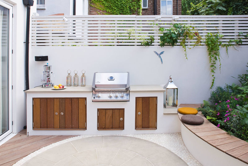 An Ibiza-inspired garden makeover featuring a vibrant mosaic wall, bespoke outdoor kitchen with BBQ, York stone dining area, handcrafted hardwood bench, and a lush herb wall. Bright pink lounge chairs and evergreen planting create a colorful, family-friendly backyard perfect for entertaining.